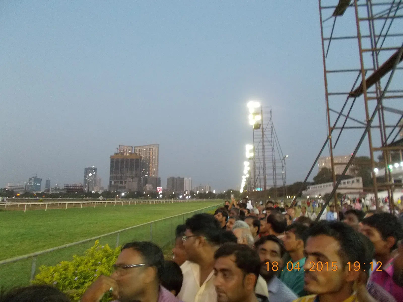 Spectators on the rail at Mahalaxmi Racecourse in Mumbai at dusk with floodlights and city skyline